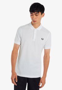 Weißes Poloshirt aus Baumwolle, mit klassischem Kragen, kurzen Ärmeln und einem kleinen schwarzen Logo auf der linken Brust.