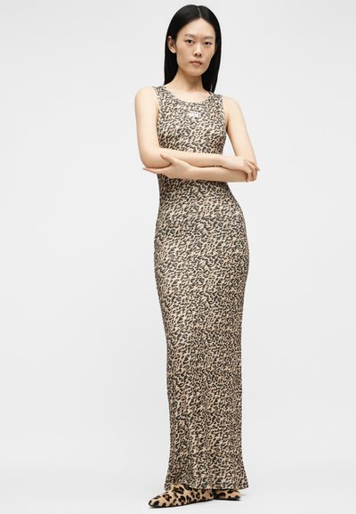 Γυναίκα που στέκεται με σταυρωμένα τα χέρια, φορώντας εφαρμοστό αμάνικο μακρύ φόρεμα με λεοπάρ σχέδιο και ταίριασμα flat παπούτσια σε μονόχρωμο φόντο.