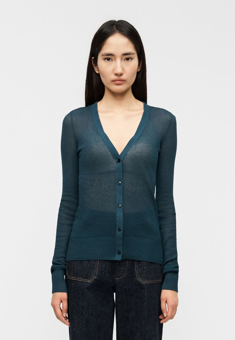 Τζελ χρώματος τιρκουάζ, ελαφρύ πλε κτό κάρντιγκαν με V-λαιμόκοψη, κλείσιμο με κουμπιά και υφασμάτινο μοτίβο στη μέση και τα μανίκια. Μακριά, εφαρμοστά μανίκια.