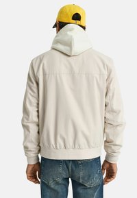 Licht beige jack met een witte capuchon, elastische zoom en manchetten. Getextureerde stof, casual ontwerp, gedragen met blauwe jeans en een gele pet.