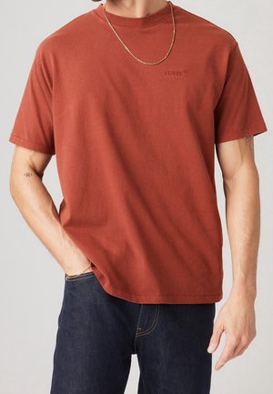 Homme portant un t-shirt Levi's couleur rouille, un jean bleu foncé, une chaîne en or autour du cou et de petits tatouages sur les deux avant-bras.