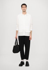 Camisa de lino blanca con botones y dos bolsillos frontales, combinada con pantalones holgados negros y mocasines negros. El modelo sostiene una bolsa tote negra.