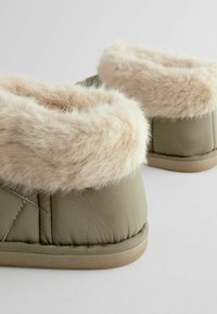 Blødt grønt hjemmesko med en plysset beige imiteret pelskant, quiltet krop og flad beige sål. Har stikninger på overdelen.