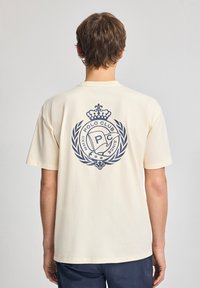 Camiseta de algodón color crema con un gran logotipo en azul marino en la parte posterior, adornado con una corona y acentos de laurel, de mangas cortas y con cuello redondo.