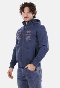 Felpa con zip di colore blu navy, realizzata in misto cotone, con tasche frontali, loghi ricamati e polsini a costine. Texture morbida con un design aderente.