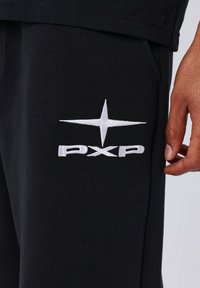 Pantalon de survêtement en coton noir avec un logo "PXP" blanc brodé et un design d'étoile sur le côté gauche. Comprend une poche latérale. Texture douce.