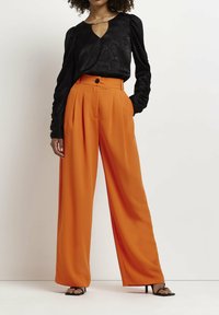 Blusa nera con texture, maniche lunghe a sbuffo e scollatura a chiave, abbinata a pantaloni arancioni a vita alta e gamba larga. Tacchi neri con laccetti.