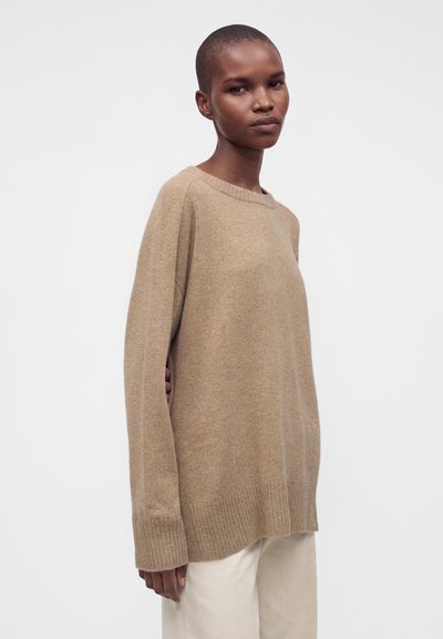Personne aux cheveux courts portant un pull beige ample et un pantalon clair se tient devant un fond clair uni.