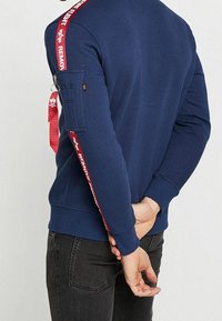 Sudadera azul marino con franjas laterales rojas y un bolsillo con cremallera en la manga, que presenta una etiqueta prominente y detalles de herrajes ajustables.