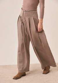 Pantalones de pierna ancha en una suave tela topo, con pliegues y cintura con cordón, combinados con botas de tobillo marrones sobre una superficie beige.