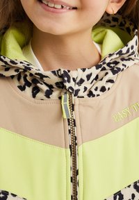 Bambino sorridente, indossa una giacca beige e verde neon con cappuccio a stampa leopardata e "EASY" ricamato sul petto vicino alla zip.