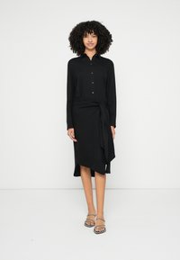 Crna haljina-košulja s dugim rukavima, ovratnikom, gumbima sprijeda i bočnim vezivanjem. Glatka tekstura, asimetrični rub, uparena sa sandalama na remenčiće.