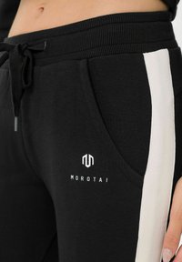 Čierne tepláky vyrobené z mäkkej tkaniny, s bielym bočným pruhom, s pásom na šnúrku a logom "MOROTAI" v oblasti prednej vrecka.