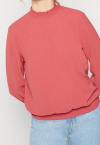 Korallin pinkki collegepaita, jossa on röyhelökaulus, pitkät hihat ja kytketty helma. Pehmeä kangas, jossa on hieman löysä istuvuus ja rento muotoilu.