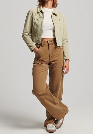 Vrouw draagt een cropped beige jas, een witte cropped top, high-waisted bruine wijde broekspijpen en witte sneakers, staat met handen in de zakken.