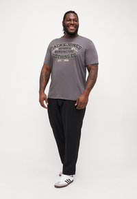 Pelēks T-krekls ar dzeltenu "Jack & Jones" logo; brīvs piegriezums. Melni bikses; mīksta auduma. Baltas kedas ar melniem un brūniem akcentiem.