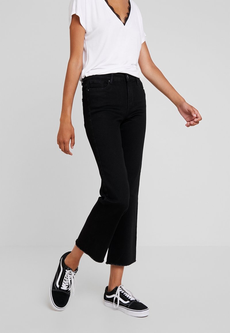 Femme portant un jean noir évasé coupé, un t-shirt blanc à col en V avec des bordures en dentelle, et des sneakers noir et blanc se tenant sur un fond neutre.