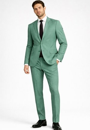 Homme debout avec une main dans la poche, portant un costume vert clair, une chemise blanche, une cravate noire et des chaussures habillées noires sur fond blanc.
