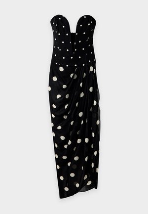 Une femme porte une robe noire bustier à pois blancs et à jupe drapée, ainsi que des sandales noires, sur un fond blanc uni.
