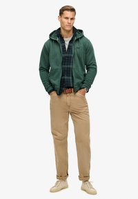 Sudadera con cremallera de color verde oscuro sobre una camisa de cuadros azul y verde, pantalones beige y zapatillas claras. Presenta un bolsillo frontal y capucha con cordón.