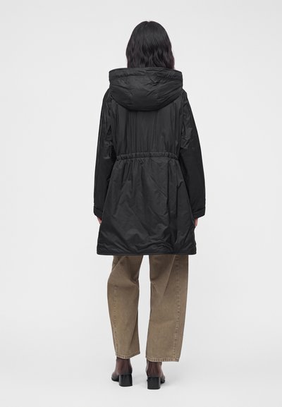 Femme portant un manteau noir à capuche avec une taille cintrée, un pantalon marron à jambes larges, et des chaussures noires à talons, debout de dos sur un fond uni.