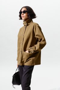 Lühikeste tumedate juustega naine, kellel on mustad päikeseprillid, pruun nööpidega jakk, mustad püksid ja must käekott käes.