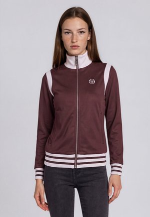 Jeune femme aux cheveux bruns et lisses portant une veste zippée bordeaux et blanche et un pantalon foncé, debout devant un fond uni.