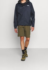 Mann trägt eine navyblaue Kapuzenjacke, olivgrüne Cargo-Shorts, schwarze Socken und olivgrüne Wanderschuhe und geht nach vorne.