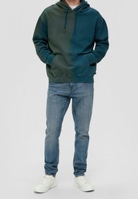 Mikina s kapucí z bavlněné směsi v tmavě zeleném přechodu, vybavená kapsou na přední straně a nastavitelnými šňůrkami. Kombinováno s modrými džínami a bílými teniskami.
