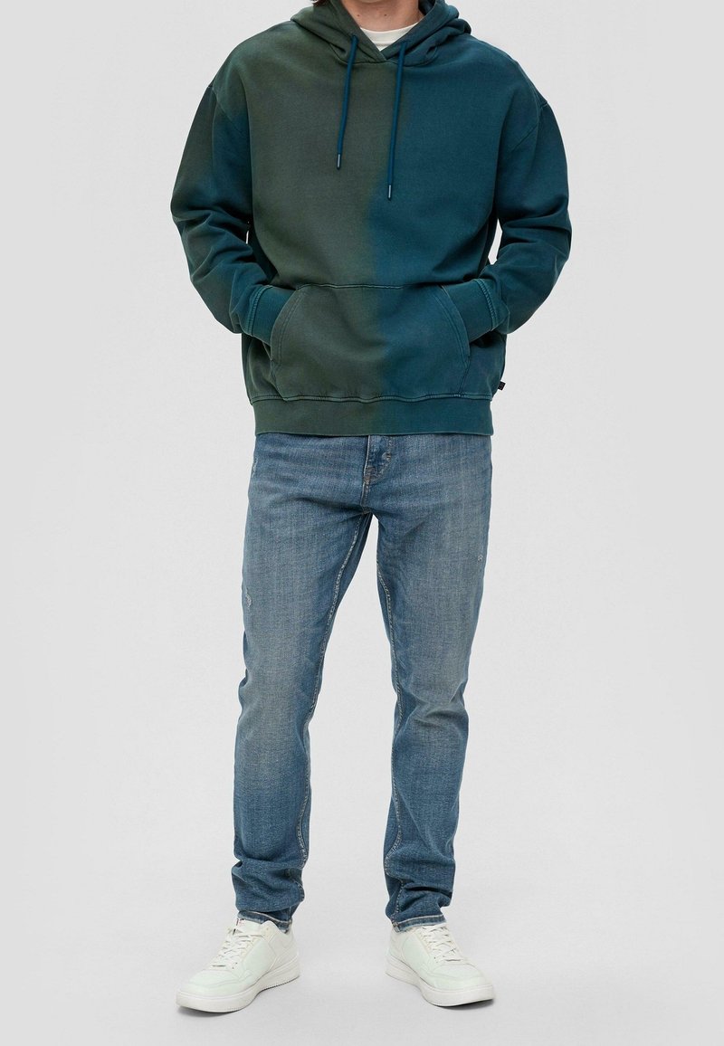 Mikina s kapucí z bavlněné směsi v tmavě zeleném přechodu, vybavená kapsou na přední straně a nastavitelnými šňůrkami. Kombinováno s modrými džínami a bílými teniskami.