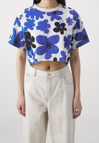 Camiseta recortada en blanco con patrones de flores azules y negras. Presenta un escote redondo y mangas cortas. Combinada con pantalones claros de cintura alta.