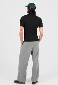 Persona che indossa una camicia nera a maniche corte, pantaloni grigi larghi, scarpe nere e un cappellino da baseball verde, in piedi con le mani in tasca girato di spalle.