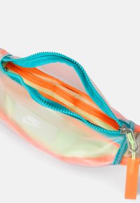 Permatomas fanny pack'as su turkio spalvos užtrauktuku, oranžiniais ir geltonais vidinės dalies skyreliais, bei maža balta Nike logotipo etikete. Sklandus paviršius.