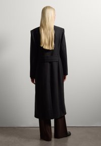 Persoon met lang blond haar draagt een lange zwarte jas en donkere wijde broek, tegen een plain witte muur staand met de rug naar de camera gekeerd.