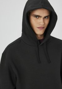 Hoodie noir avec une grande capuche, des cordons de serrage et une texture douce. Présente une coupe décontractée et des détails de couture minimalistes sur les épaules.