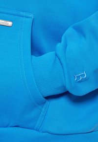 Niebieski sweatshirt z prążkowanymi mankietami, kieszenią z przodu i detalem z logo. Gładka faktura i jednolity kolor w całym ubraniu.
