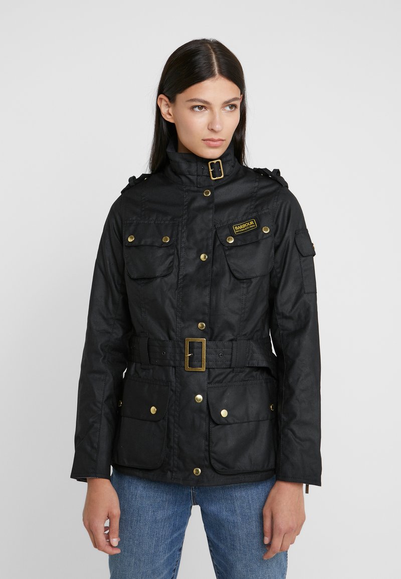 Black waterproof jacket with a high collar, gold hardware, four pockets, a belted waist, and shoulder detailing. Smooth texture and practical design.
