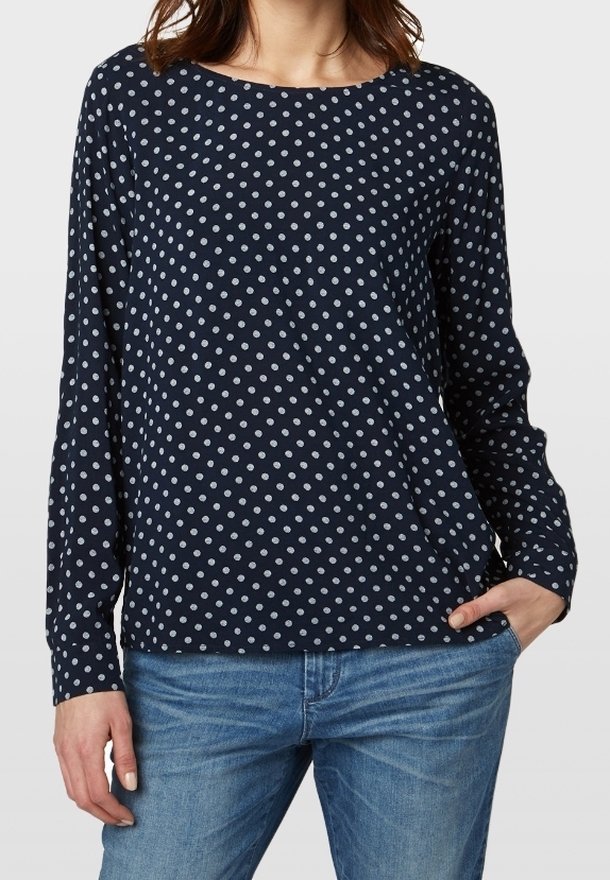 Blouse à manches longues navy avec des pois blancs, décolleté bateau et coupe décontractée. Tissu doux avec une texture lisse. Associée à un jean bleu clair.