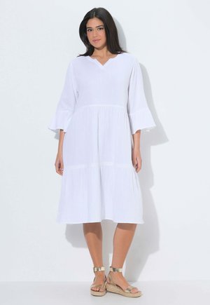 Kobieta o ciemnych włosach ubrana w białą sukienkę do kolan z dzwonkowatymi rękawami i złote sandały na koturnie z paskiem wokół kostki, stojąca na białym tle.