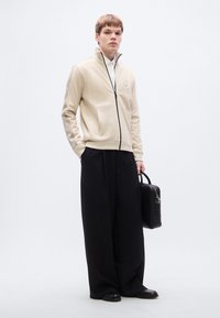 Μπεζ μπουφάν με φερμουάρ και πλαϊνές τσέπες, μαύρο φερμουάρ και λογότυπο. Συνδυάζεται με άνετο μαύρο παντελόνι και κρατάει μια μαύρη ορθογώνια τσάντα.