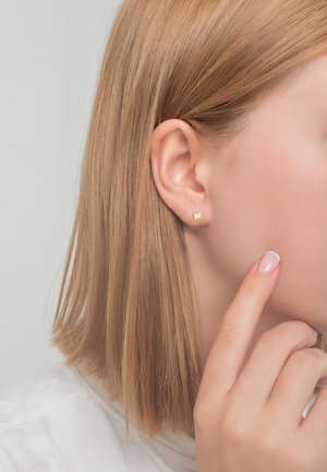 Goldene Steckerohrringe mit strukturiertem Design, getragen an einem hellhäutigen Ohr. Die Haare sind glatt und schulterlang, der Hintergrund ist neutral.