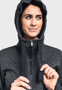 Chaqueta con cremallera de patrón gris y capucha, con detalles en negro y una textura suave. El modelo sostiene los cordones de la capucha con ambas manos.