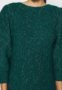 Maglione lavorato a maglia teal con filato testurizzato e scintillante. Presenta un collo a giro e maniche a tre quarti. Sono visibili dei motivi a diamante.