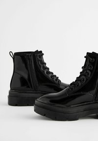 Bottes chelsea en cuir verni noir avec une semelle épaisse, fermeture à lacets à l'avant et zip sur le côté. Présente une semelle noire texturée et une languette ajustable.