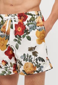 Costume da bagno floreali con sfondo bianco, grandi rose rosse, gialle e arancioni, foglie verdi e una vita con coulisse.