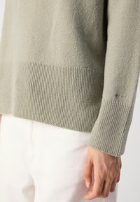Close-up of a person's hand beside the hem and cuff of a light gray knit sweater, with white pants visible in the background.