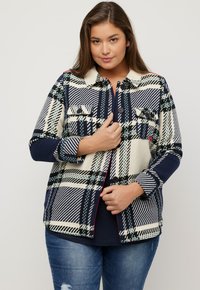 Giacca a camicia a quadri in blu navy, crema e verde; presenta una chiusura con bottoni, due tasche sul petto e un colletto; tessuto morbido.