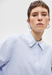 Γυναίκα με κοντά καστανά μαλλιά, φορώντας ασημένια σκουλαρίκια κρίκους και ένα ανοιχτό μπλε ριγέ πουκάμισο με λευκό δαντελένιο γιακά, μπροστά σε λευκό φόντο.