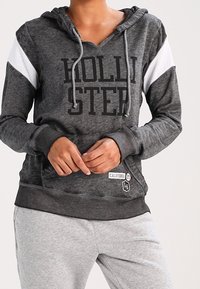 Šedá mikina s kapucí s černým nápisem "HOLLISTER", bílošedé pruhované rukávy, přední kapsa a nášivka Kalifornie. Měkký materiál a volný střih.