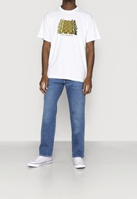 Bílé bavlněné tričko s vícebarevným grafickým potiskem na přední straně. Kombinováno s modrými džínami a světle fialovými teniskami.